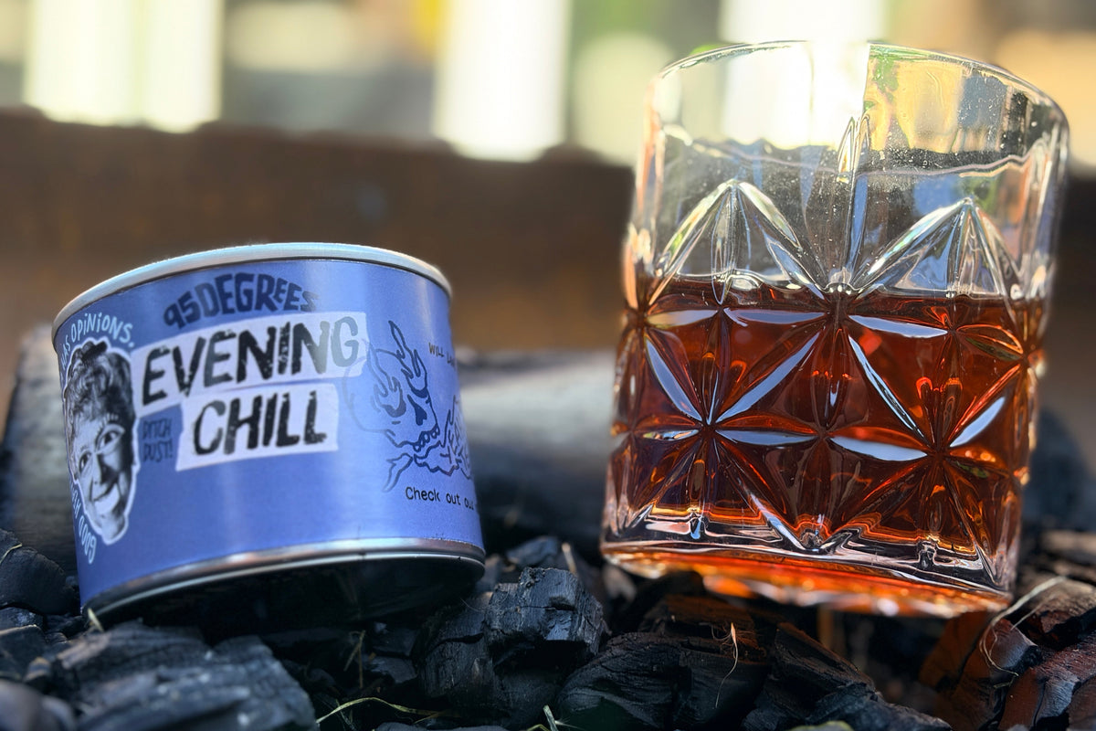 Close-up lifestyle shot of a blue 95 Degrees Evening Chill Lapsang Souchong loose leaf tea tin and a crystal glass of freshly brewed dark tea, placed on a bed of dark charcoal with a soft, blurred background.