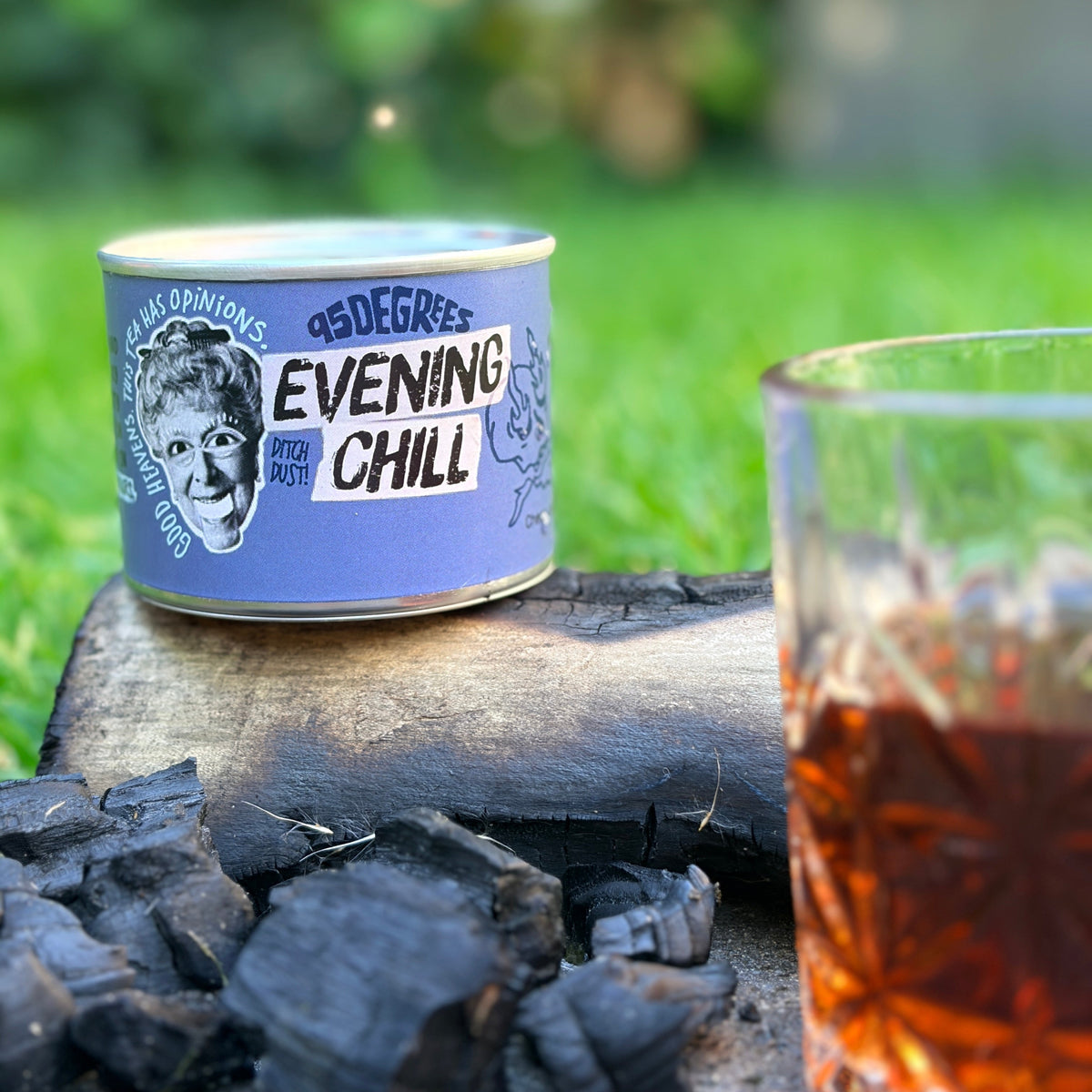 A lifestyle shot of a blue 95 Degrees Evening Chill lapsang souchong tea tin resting on charred charcoal logs in a garden, with a crystal glass of amber-colored brewed tea in the blurred foreground.