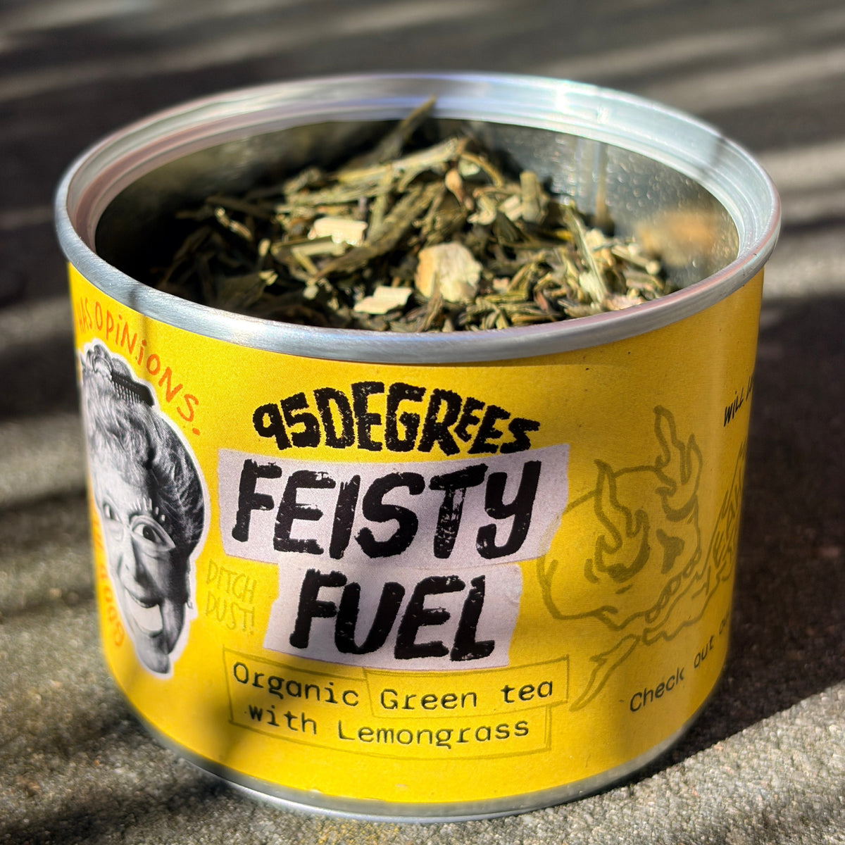 A close-up of an open round tea tin with a bright yellow label. The label reads '95 Degrees FEISTY FUEL - Organic Green tea with Lemongrass'. It features a black-and-white illustration of a face on the left and a flame graphic on the right. The tin is filled with dried green tea leaves and lemongrass pieces.
