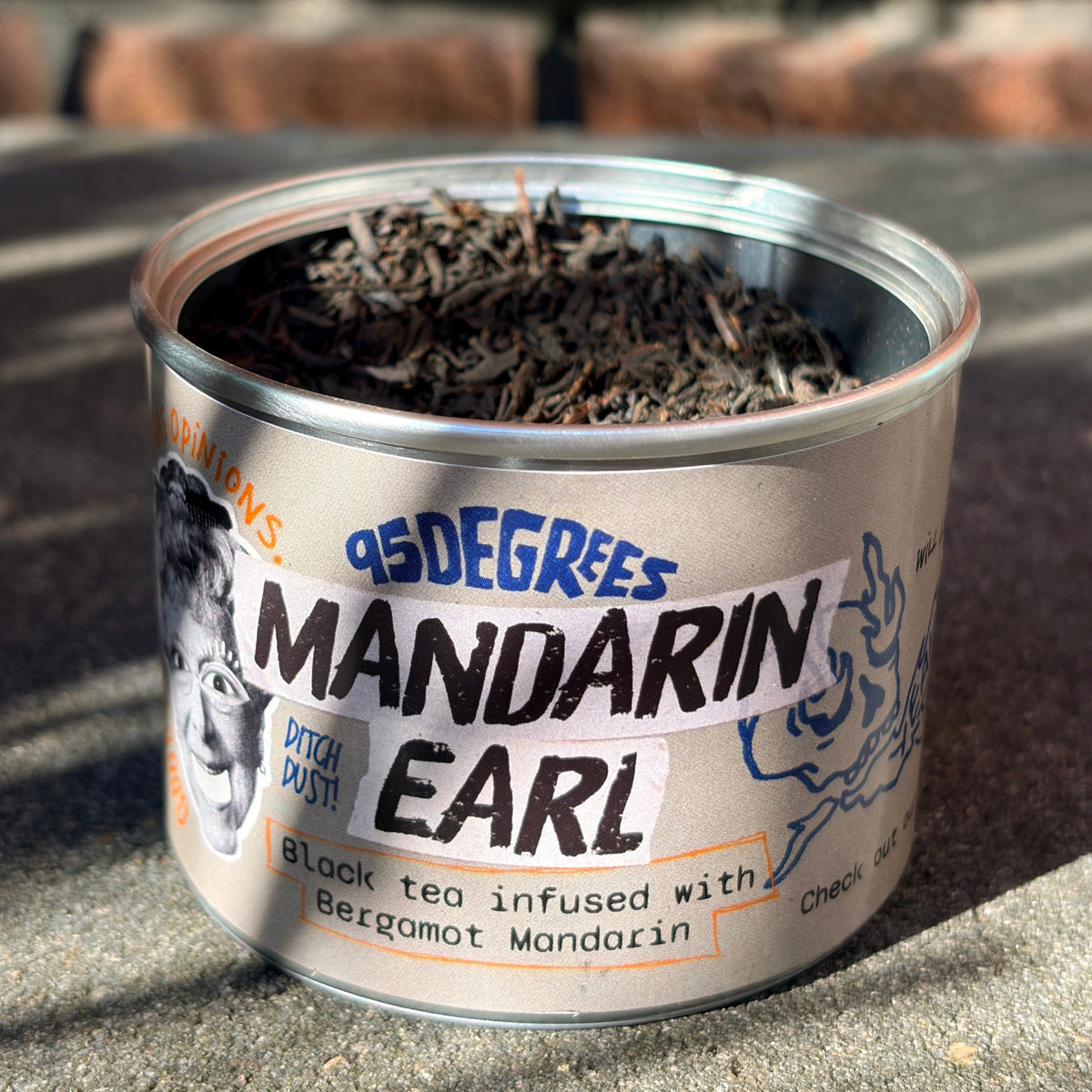 
A close-up of an open round tea tin with a silver label. The label reads '95 Degrees MANDARIN EARL - Black tea infused with Bergamot Mandarin'. It features a black-and-white illustration of a face on the left and a rose graphic on the right. The tin is filled with dark, loose-leaf black tea.

