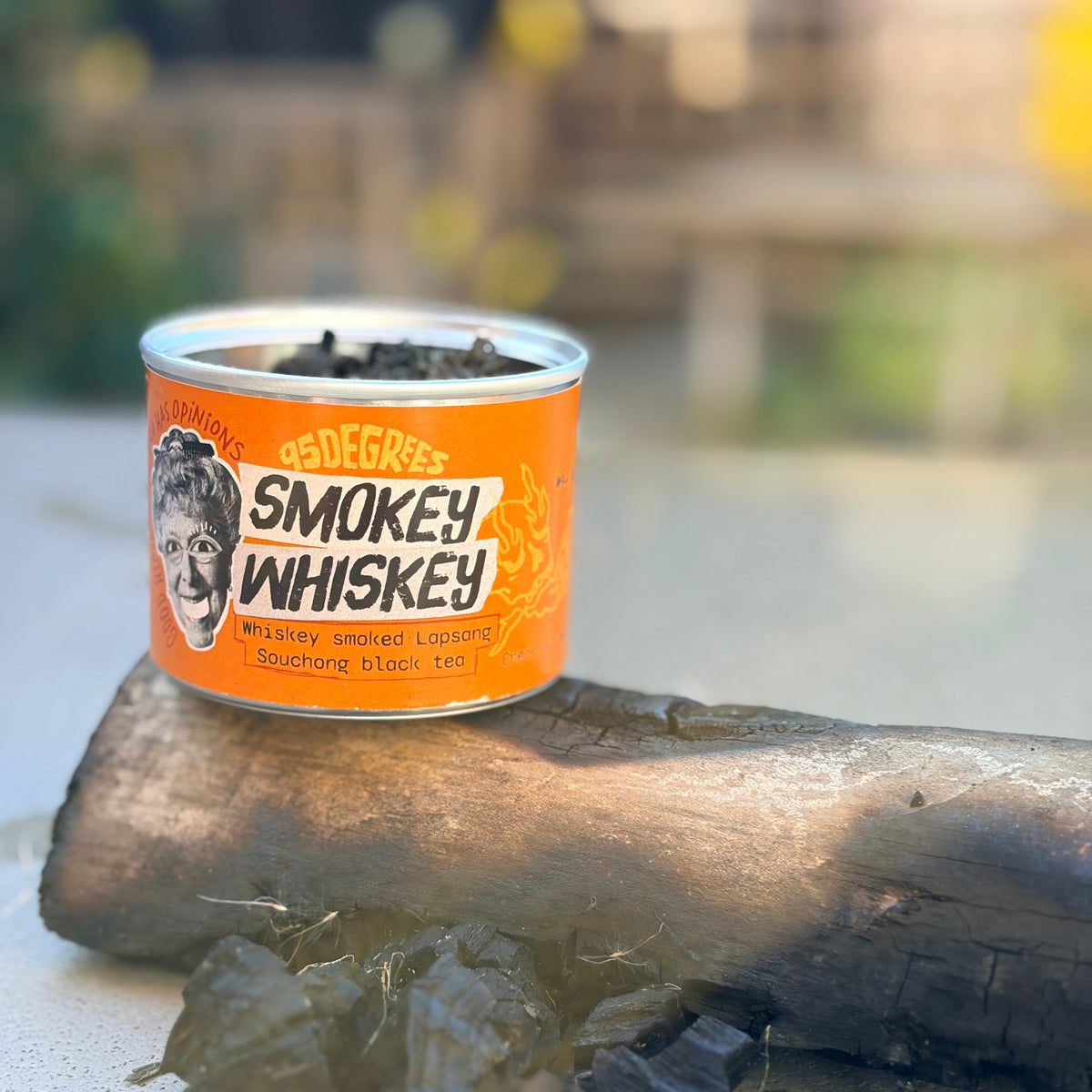 An open orange tin can of 95degrees rocks Smokey Whiskey black tea balanced on a burnt log in natural outdoor lighting.
